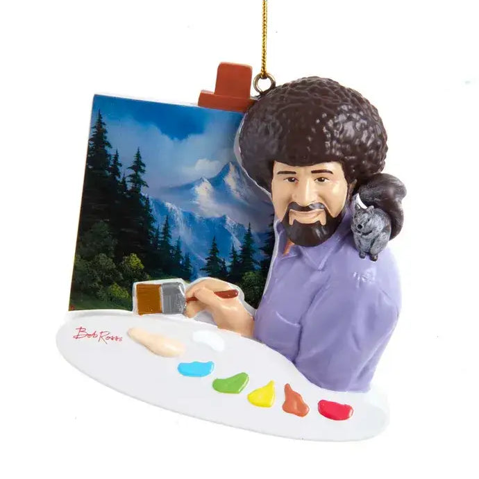 Ornament of a retro-style Bob Ross holding a paint palette and brush, sitting in front of a mountain landscape canvas with a small animal on his shoulder.