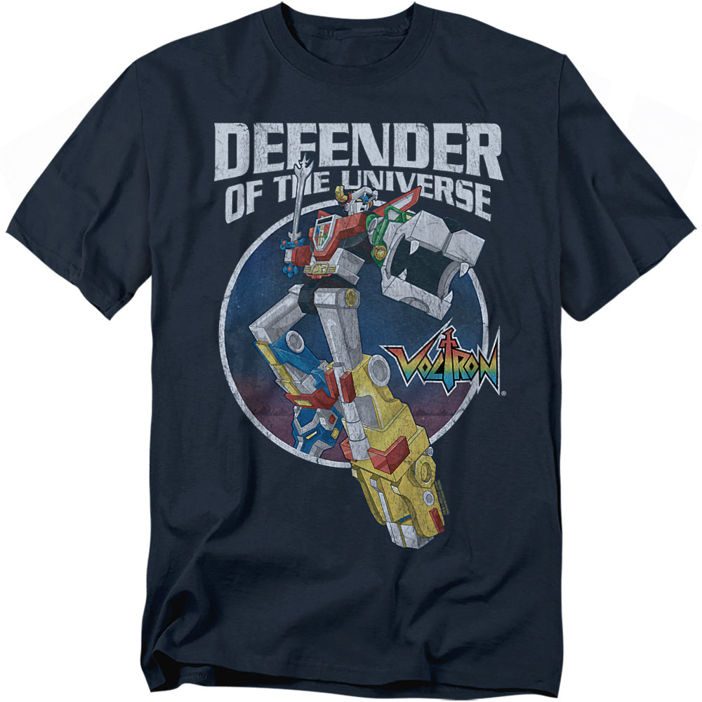 A navy blue t-shirt featuring Voltron, the giant robot made of five lion mechas, standing in a dynamic pose with one arm raised and the other extended forward. Behind him is a circular galaxy-style background. Above the design, bold white text reads “DEFENDER OF THE UNIVERSE,” with the colorful Voltron logo to the right.