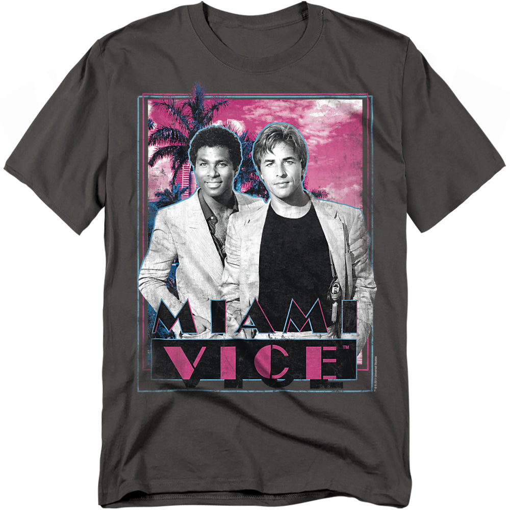 Charcoal t-shirt featuring Crockett and Tubbs from Miami Vice standing in front of palm trees with a pink neon sunset and bold logo.