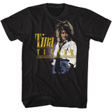 Black T-shirt featuring a distressed vintage-style photo of Tina Turner wearing a white blouse and high-waisted pants, with bold yellow “Tina Turner” text and horizontal accent lines.